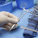 Technician picks up needle holder and suture from sterile field in operating room.